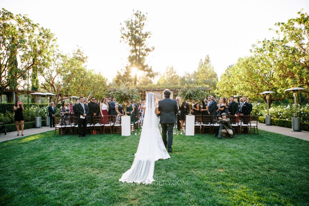 A bride, wearing a long white dress and veil, walks down an outdoor aisle at Rancho Bernardo Inn with a man, possibly her father, towards an altar. Friends and family stand on either side, surrounded by greenery and trees, capturing the moment with cameras.