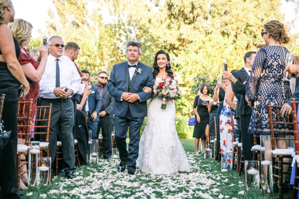 A bride in a white dress walks down an outdoor aisle at Rancho Bernardo Inn covered with white rose petals, accompanied by an older man in a suit. Attendees seated on both sides of the aisle watch and take photos. Trees and greenery are in the background.