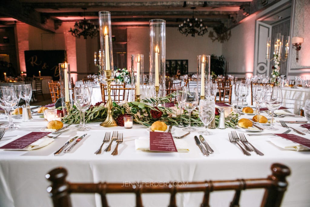 A beautifully set dining table for a Rancho Bernardo Inn wedding, adorned with tall candles in glass holders, greenery garlands, and place settings with wine glasses, silverware, and napkins with menus. The backdrop includes elegant lighting and floral arrangements—perfect for getting started as a photographer.