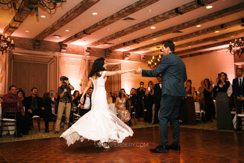 A bride in a white wedding dress twirls on the dance floor, holding hands with the groom in a suit. Guests surround the couple, smiling and clapping. The reception hall is elegantly decorated with chandeliers and wooden ceiling beams.