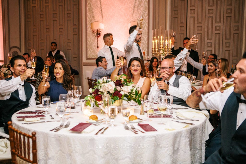 A group of elegantly dressed people sitting around a round dining table raise their glasses in a toast at what appears to be a wedding reception. The table is decorated with flowers and candles, and the room features sophisticated lighting and decor.