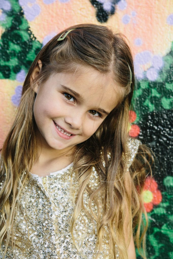 A young girl with long hair smiles in front of a colorful, textured background. She wears a sparkly, sequined top and a small hair clip, exuding a joyful and playful demeanor.