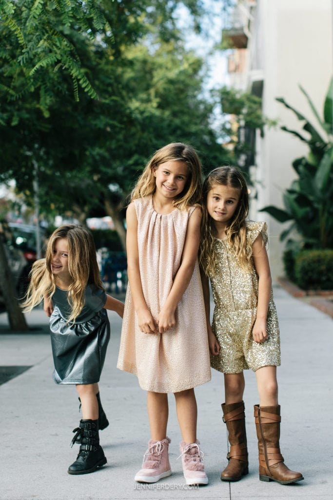 Three young girls stand outdoors on a sidewalk. One wears a gray dress and black boots, playfully bending. The middle girl is in a light pink dress with pink sneakers. The third girl wears a sparkly gold dress with brown boots. Trees and buildings are behind them.