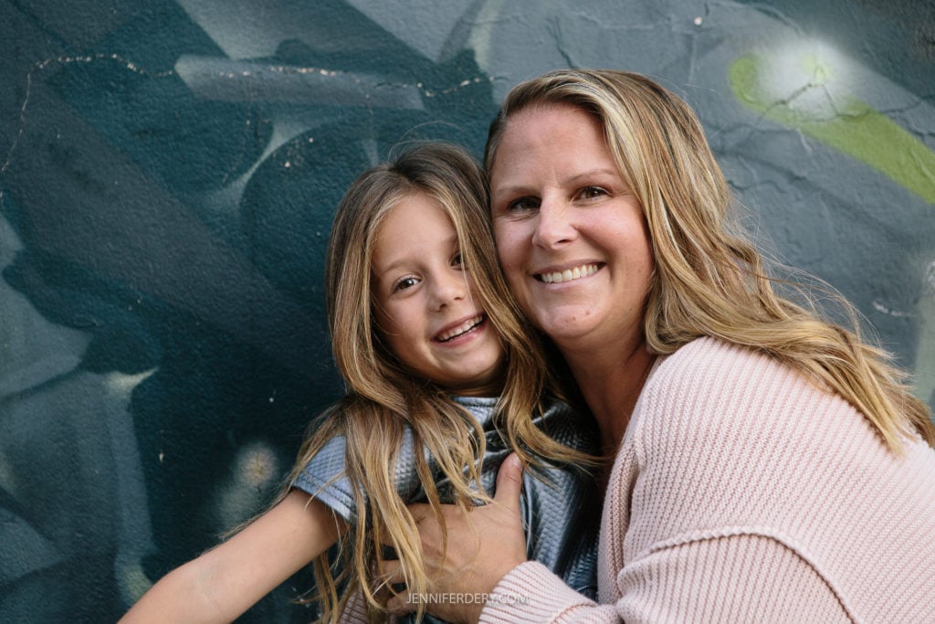 A woman and a young girl smile warmly at the camera, embracing in front of a textured, graffiti-style wall. The woman has long blond hair and wears a light sweater, while the girl has long hair and wears a striped outfit.
