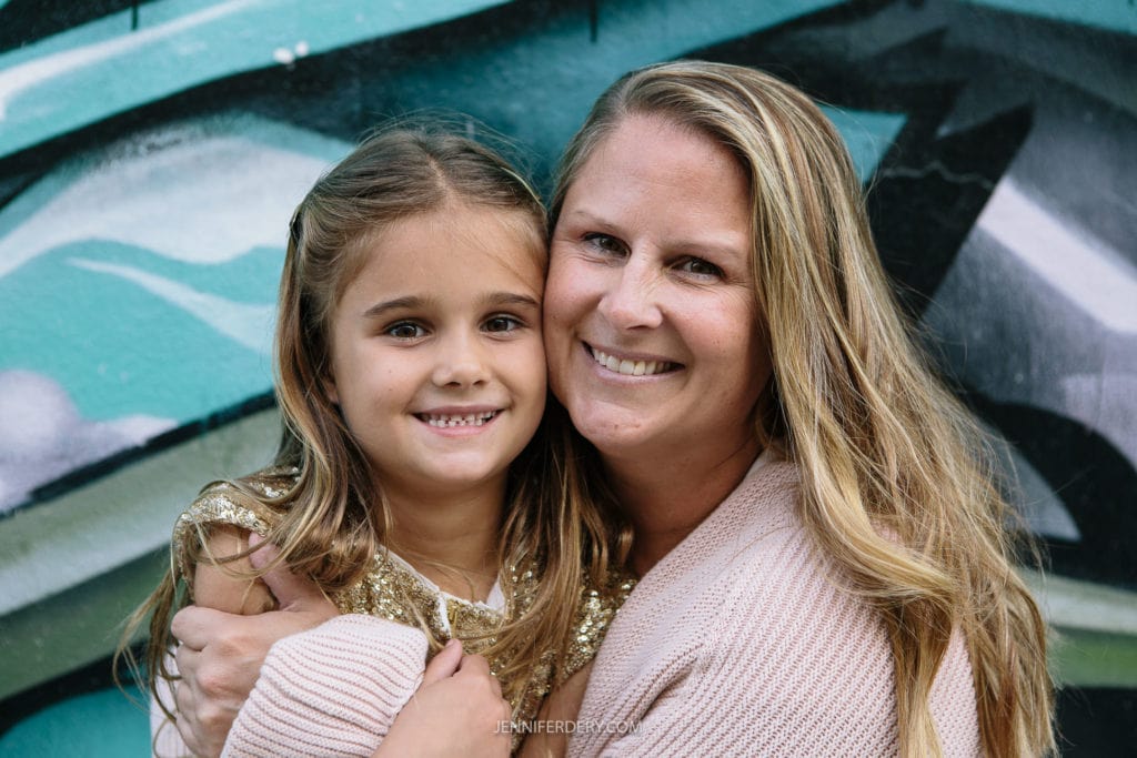 A woman and a young girl smile while hugging in front of a colorful mural. The girl wears a gold sequined top; the woman wears a light knitted sweater. Both have long hair and share a joyful expression.