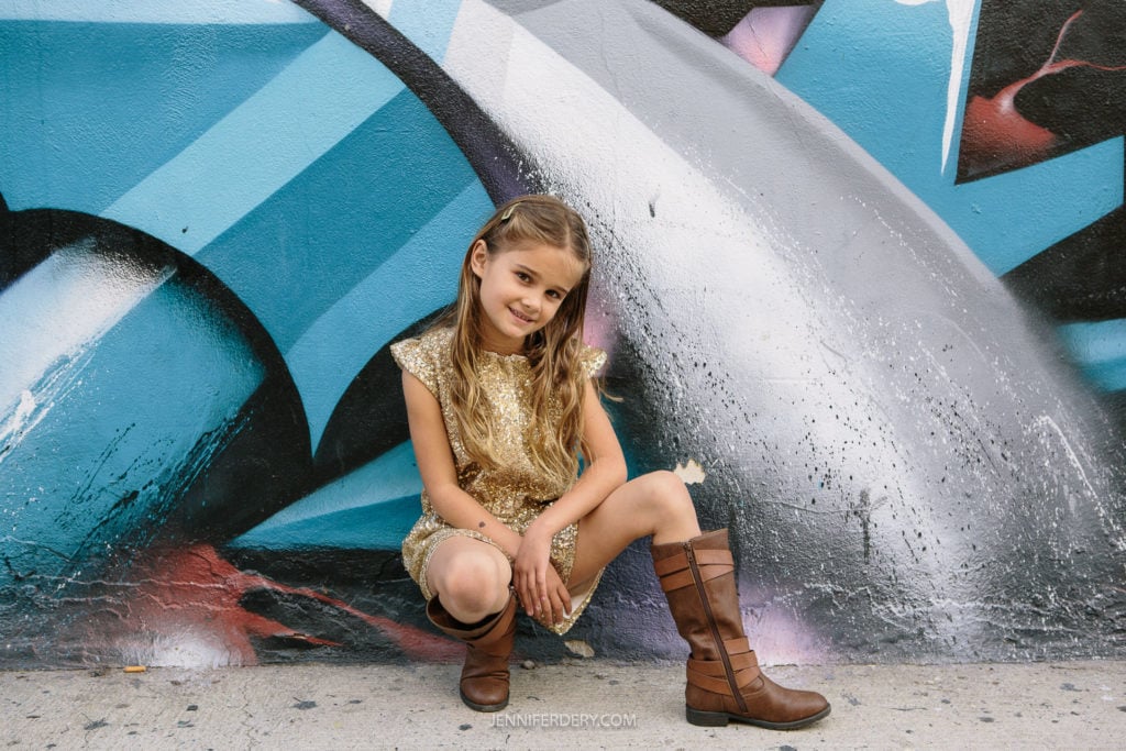 A young girl crouches in front of a colorful graffiti wall. She wears a sparkly gold dress and brown boots, gazing at the camera with a slight smile. Her hair is styled in two braids.
