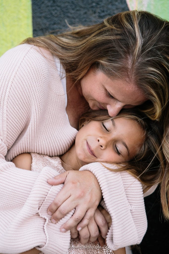 A woman in a light pink sweater lovingly holds a young girl in a tender embrace. The girl has her eyes closed, wearing a lace outfit. The background has colorful, abstract patterns.