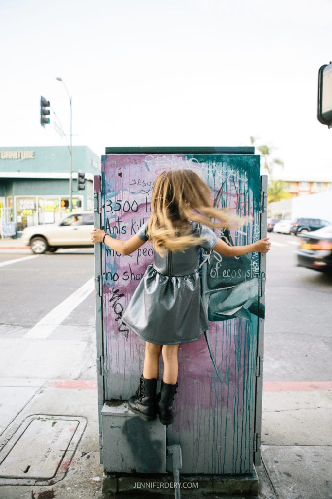A child with long hair, wearing a silver dress and black boots, is climbing on a painted utility box at a street corner. The street is busy with moving cars and a furniture store is visible in the background.