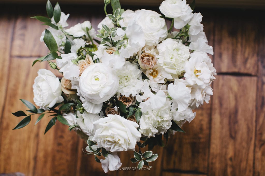 A bouquet of white and cream-colored flowers arranged with green foliage on a wooden surface. The flowers include roses, peonies, and ranunculus, creating a lush and elegant display reminiscent of Rancho Bernardo Inn Wedding Photos. The name "JENNIFERDERY.COM" is visible at the bottom center of the image.