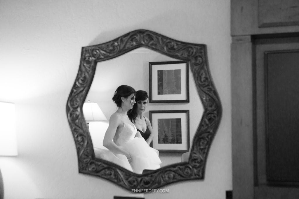 A black and white photo reflecting in a decorative mirror captures a bride being assisted with her dress by her sister. The background showcases framed artwork on the wall and a lit lamp, embodying the elegance of Rancho Bernardo Inn Photos. The photo bears the website name JENNIFERDRY.COM.