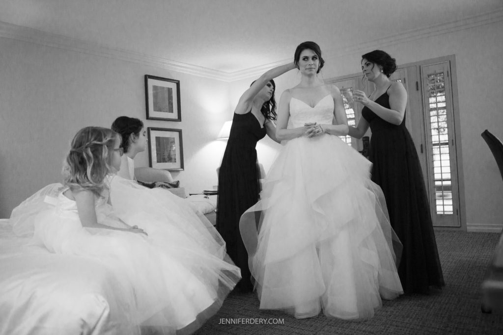 A bride stands in a room in her wedding dress as three women assist her. Two women in dark dresses arrange her hair and veil, while a young girl in a white dress sits on a bed. The scene exudes pre-wedding preparation and focus, capturing the intimate moment of Rancho Bernardo Inn wedding photos.