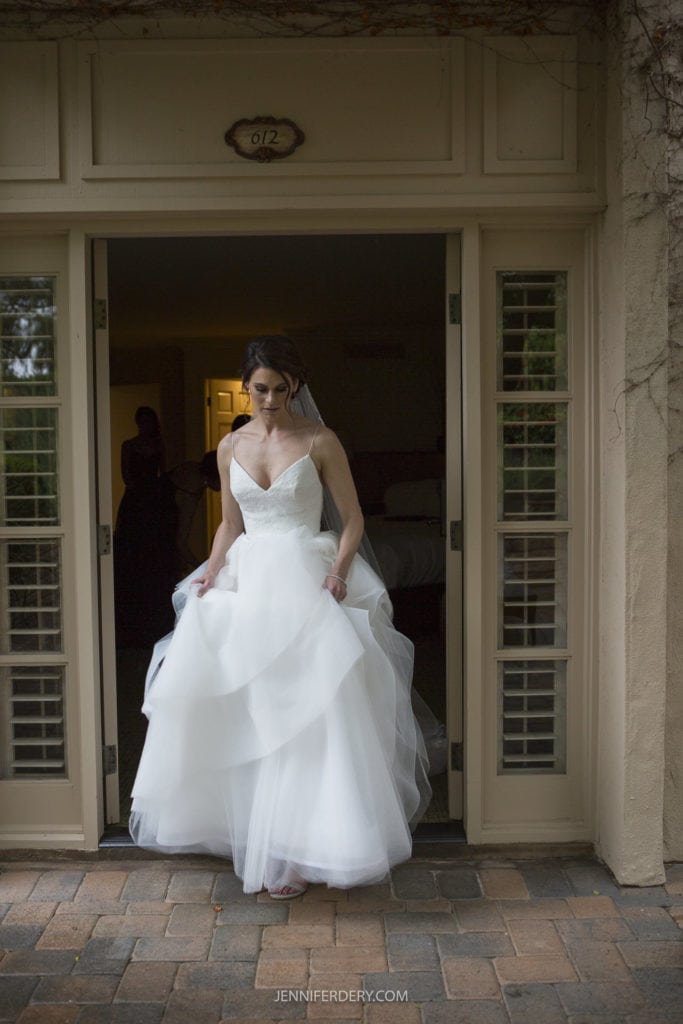 A bride in a white wedding dress and veil stands in a doorway with glass panels on both sides, appearing to step outside onto a brick path. The background reveals a partially visible room outside Rancho Bernardo Inn. The image has soft lighting, creating a serene atmosphere perfect for wedding photos.