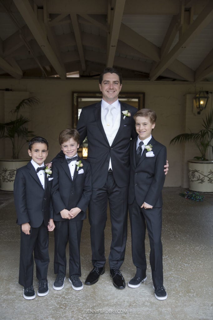 A man and three young boys, all wearing black suits, white dress shirts, and black bow ties, stand together smiling. The man has his arms around two of the boys. They are outdoors at Rancho Bernardo Inn with some plants and a window in the background; a perfect setting for wedding photos.