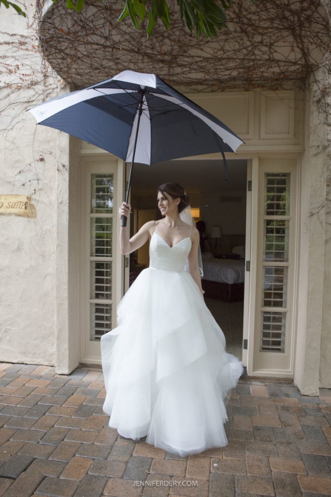 A bride in a white wedding dress stands under a dark blue and white umbrella outside of Rancho Bernardo Inn. In front of a building with double doors and ivy growing on the wall, she poses gracefully. The ground is paved with bricks, and greenery peeks in at the top of the image.