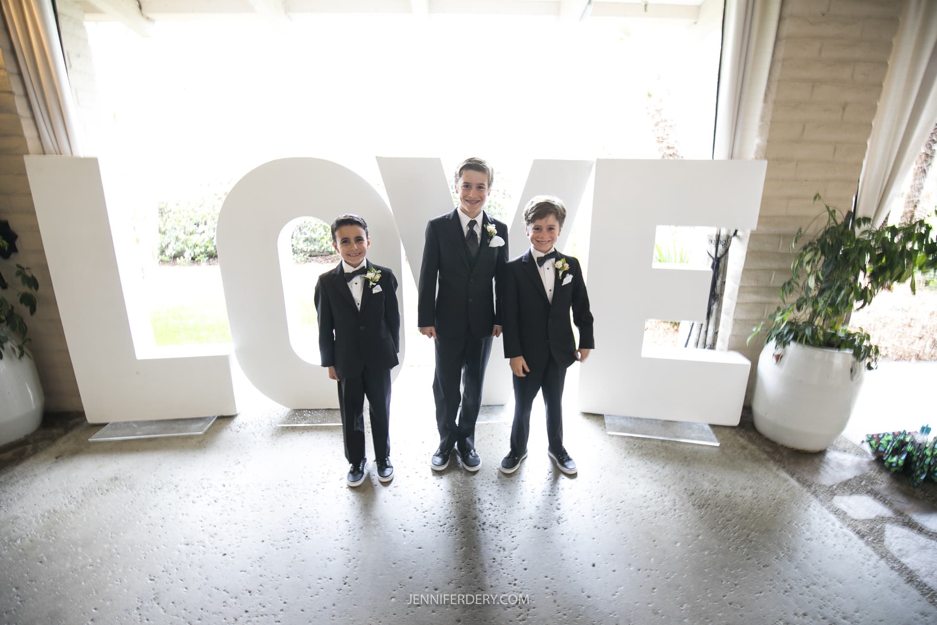 Three boys wearing black suits with boutonnieres stand in front of large, white letters spelling "LOVE." They are smiling and posing for the photo in an event venue with light spilling in from the background. Large white flower pots are visible on either side, a perfect touch for Founders Chapel wedding photos.