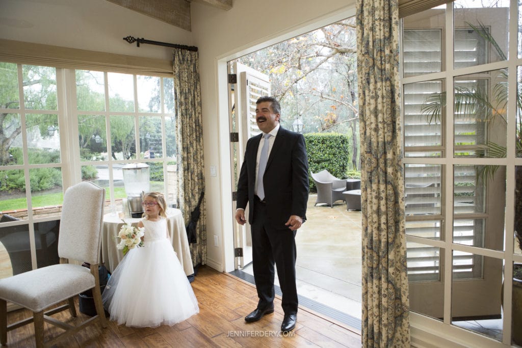 Father of the bride stands smiling in the doorway of a room with open French doors, bringing in natural light. To his left, a young girl in a white dress, presumably a flower girl, holds a small bouquet and smiles. The room is elegantly decorated with floral curtains, capturing perfect Founders Chapel wedding photos.