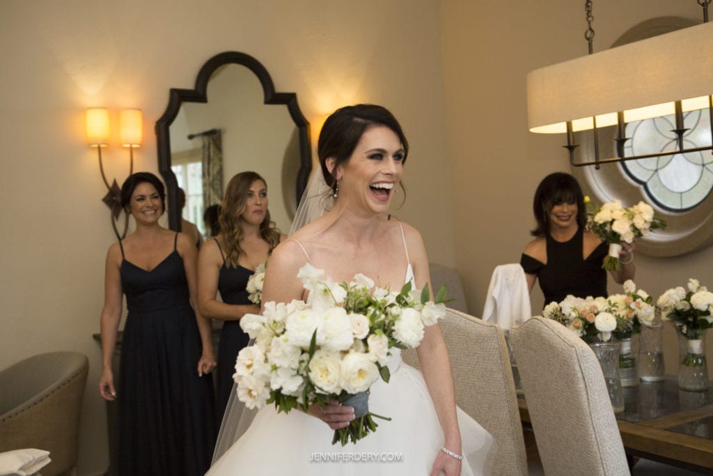 A bride in a white dress, holding a bouquet of white flowers, smiles broadly and looks to the side. Behind her, three bridesmaids in dark dresses also smile. The room at Founders Chapel has warm lighting, mirrors, and a chandelier with a circular window in the background.