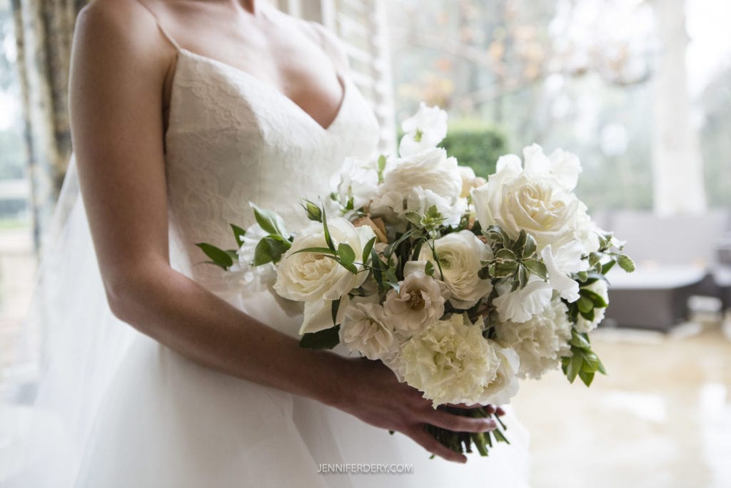 A bride in a white wedding dress holds a large bouquet of white roses and greenery. The background of the Founders Chapel wedding photo is softly blurred with natural light filtering through a window, highlighting the delicate beauty of the flowers.