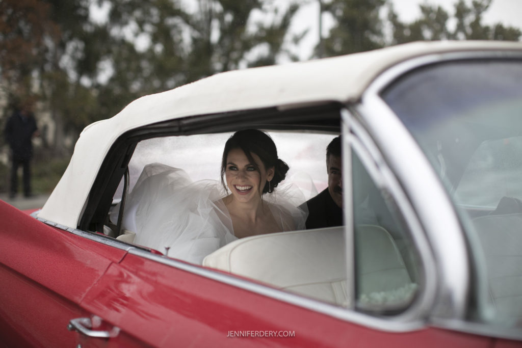 A bride in a white wedding dress smiles while sitting in the back seat of a red convertible car with a white roof, capturing the joy of Founders Chapel Wedding Photos. Trees and greenery are visible through the car windows.