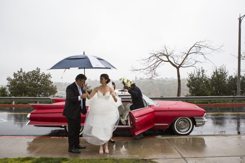 A bride in a white dress exits a vintage red car on a rainy day at Founders Chapel, assisted by a man holding an umbrella. Another person holding a bouquet is seen inside the car. The background features overcast skies and a leafless tree, perfect for capturing timeless wedding photos.