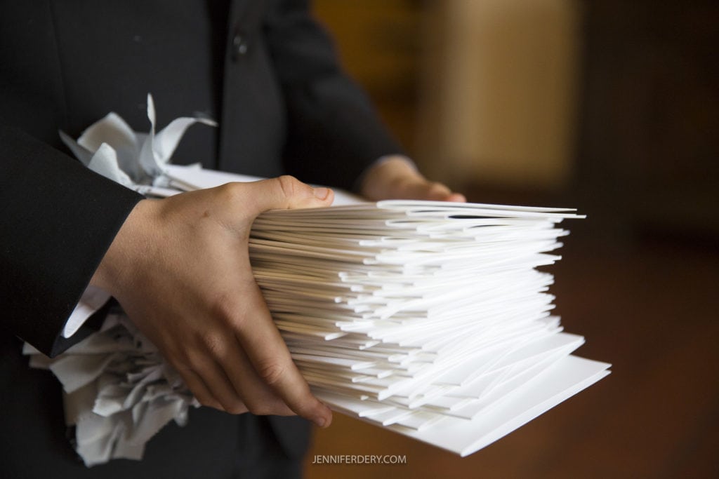 Person holding a large stack of white envelopes, possibly Founders Chapel wedding photos. They are dressed in dark clothing, with an indoor background in soft focus, making the envelopes the main focal point of the image.