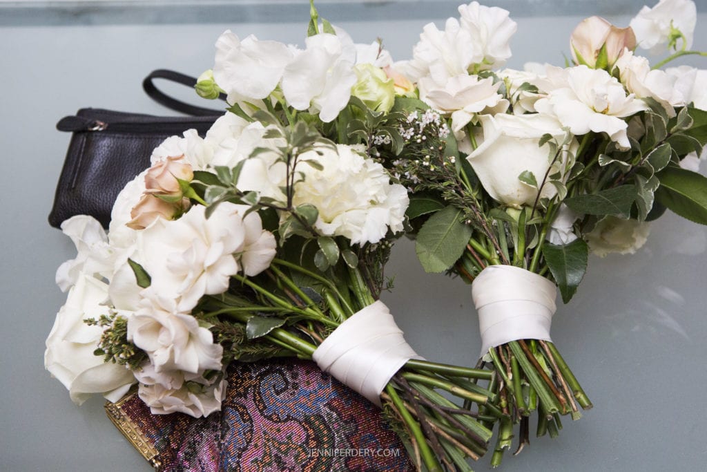 Close-up of two bridal bouquets with white and pale pink flowers, wrapped in white ribbon, placed on a glass surface. A black clutch and a colorful, patterned wallet are partially visible beside the bouquets. Captured among the Founders Chapel Wedding Photos, these details evoke timeless elegance.