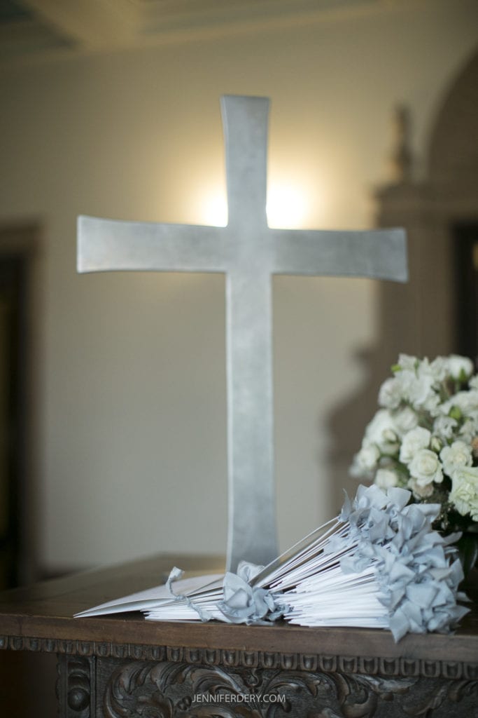 A table decorated with a large metallic cross standing upright, surrounded by white flowers and folded papers with grey ribbons, captures the essence of Founders Chapel Wedding Photos. The background features soft lighting, enhancing the serene and solemn atmosphere.