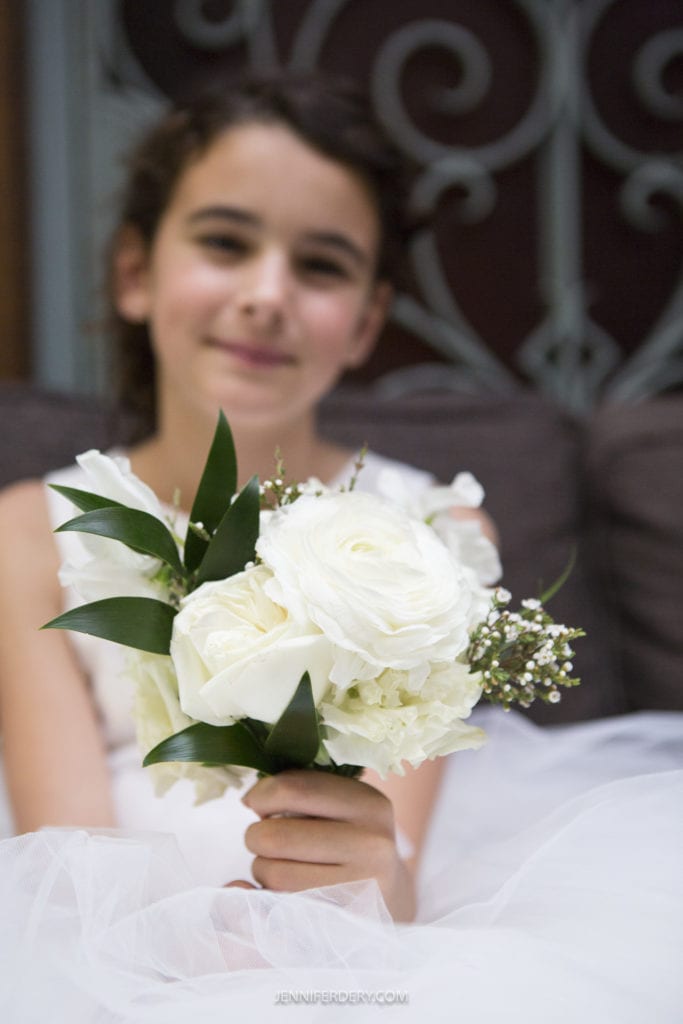 A young girl with dark hair, wearing a white dress, holds a bouquet of white roses and greenery. She is seated indoors on a cushion, with an ornate metal backdrop in the background—an exquisite moment captured as part of the Founders Chapel wedding photos. The focus is on the bouquet, her face slightly blurred in the background.