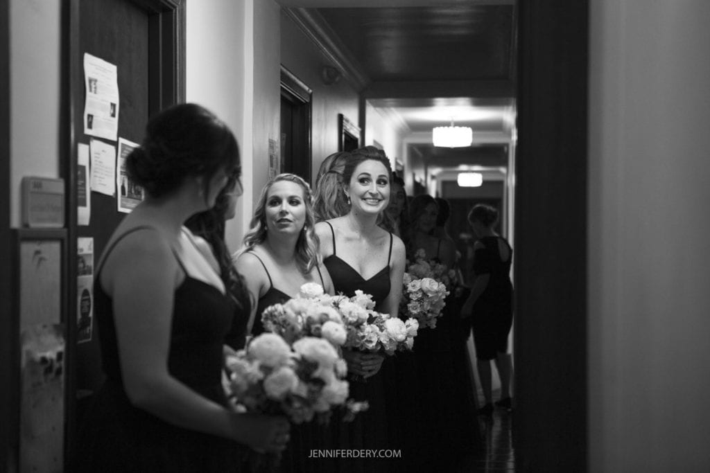 A group of women, each holding bouquets of flowers, stand in a dimly lit hallway. Dressed in matching sleeveless gowns, they engage in conversation, smiling and laughing. The scene feels intimate and joyful—an ideal moment for Founders Chapel wedding photos, capturing the essence of cherished bridesmaids.