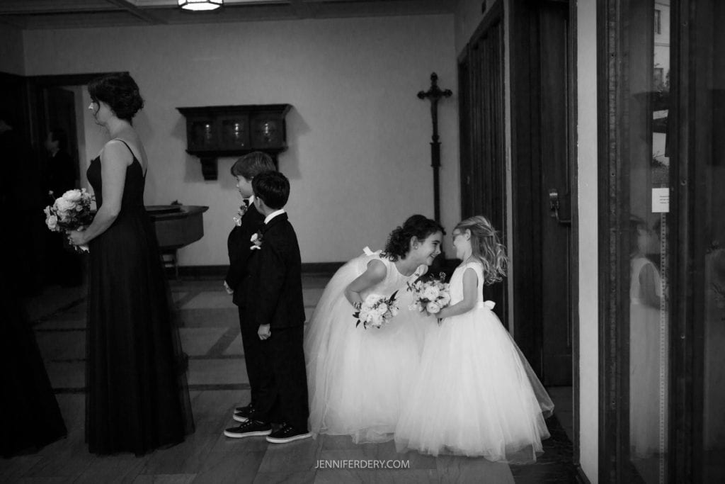 A black and white photo at a Founders Chapel wedding shows two young flower girls in white dresses, chatting and smiling near a closed door. A bridesmaid in a long dark dress stands to the left, while a young ring bearer and a boy in suits stand nearby holding bouquets.