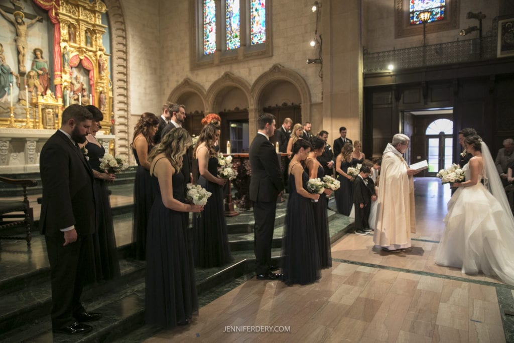 A wedding ceremony inside Founders Chapel with stained glass windows. The bride in a white gown and the groom in a tuxedo stand before a priest, while the bridal party wearing navy blue dresses and black suits stand on either side. Family and friends are seated in the pews, capturing timeless Wedding Photos.