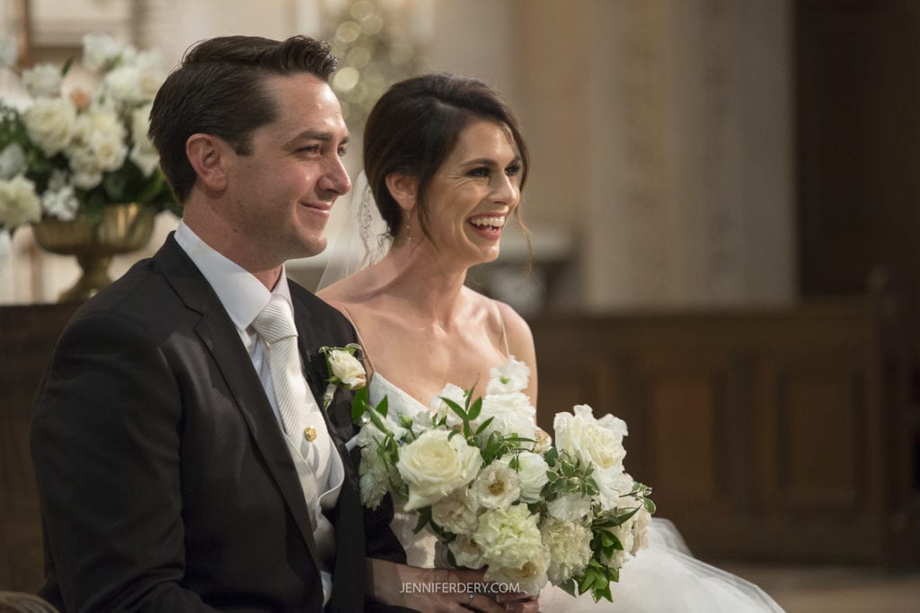A bride and groom sit side by side, smiling and holding a bouquet of white flowers. The groom is dressed in a black suit with a white boutonniere, while the bride dazzles in a white gown with an elegant updo. Captured at their wedding ceremony at Founders Chapel, the scene is adorned with beautiful floral decorations.