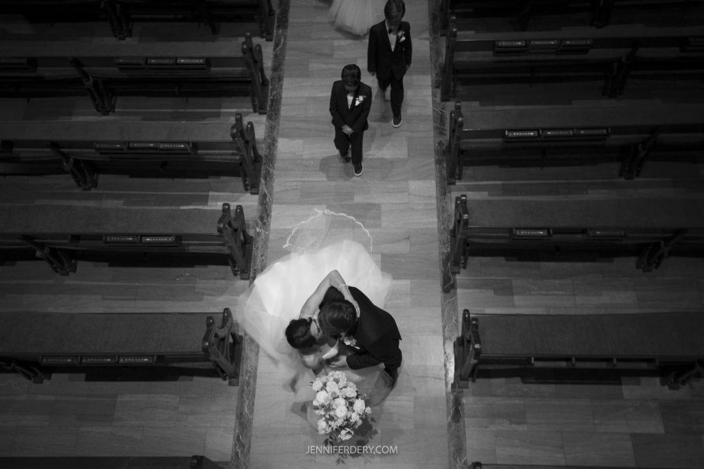 A black-and-white image captures a Founders Chapel wedding ceremony from an aerial view. The bride and groom are sharing a kiss, with the bride holding a bouquet. Two flower girls are walking down the aisle behind them. The church pews are symmetrically arranged on either side.