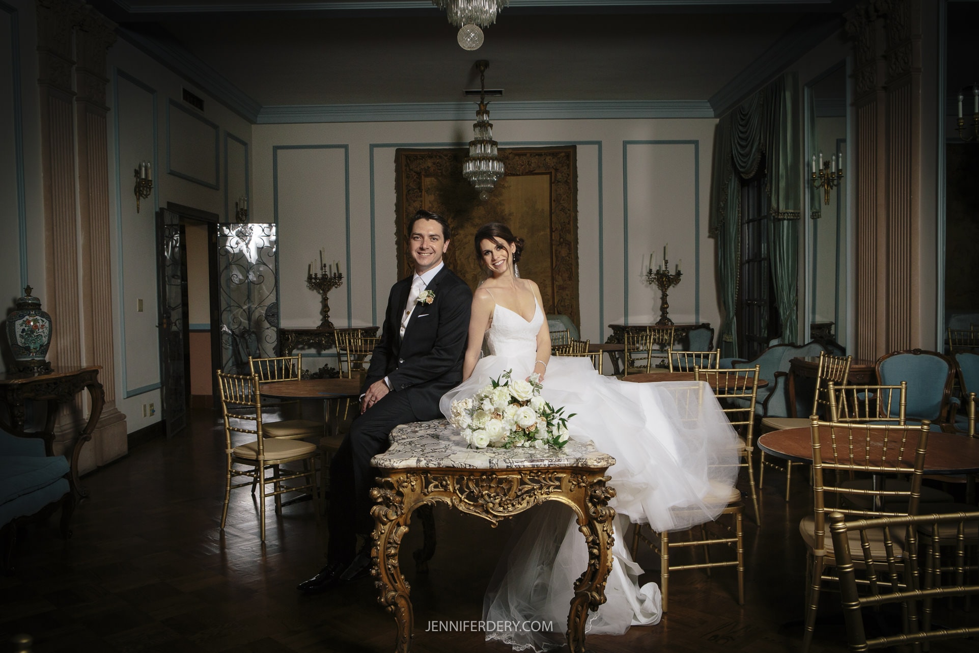 A bride and groom sit smiling on the edge of an ornate table in a lavishly decorated room with blue and gold accents, chandeliers, and elegant furniture. The bride wears a white wedding dress, while the groom sports a black tuxedo with a bow tie. These cherished Founders Chapel Wedding Photos capture their joy perfectly.
