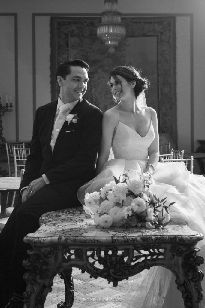 A bride and groom sit closely together on a decorated table, smiling at each other during their Founders Chapel wedding. The bride wears a white dress and the groom dons a tuxedo. A bouquet of flowers rests on the table in front of them, with elegant decor and a chandelier in the background.