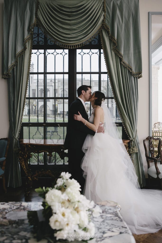 A bride and groom share a kiss in front of a large window with elegant green drapes. The bride wears a white wedding dress with a flowing veil, and the groom is in a black suit. A bouquet of white flowers on the table adds charm to this beautiful moment captured in their Founders Chapel Wedding Photos.