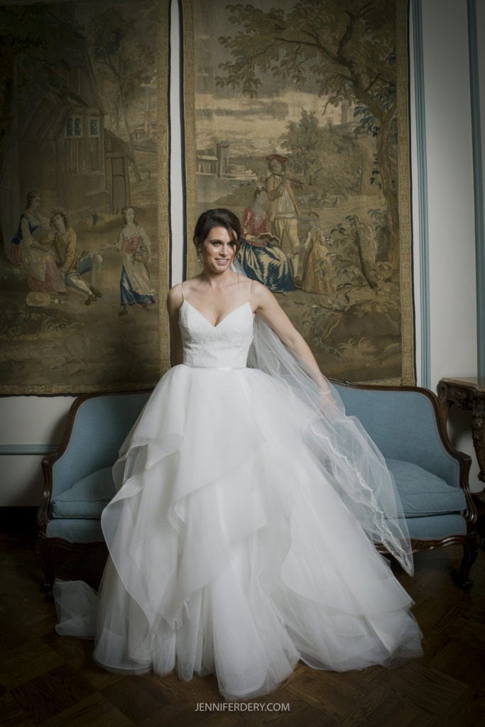 A bride in a white, strapless wedding gown with a layered, voluminous skirt stands with one hand on her waist and the other holding her veil. She poses in front of a vintage tapestry and a blue couch, smiling with a classical backdrop. This stunning moment is part of Jennifer Dery's Founders Chapel Wedding Photos.