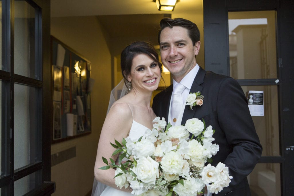A happy couple on their wedding day stands in a doorway at Founders Chapel. The bride, in a white dress and veil, holds a bouquet of white flowers and greenery. She has dark hair and smiles brightly. The groom, in a black suit and tie, stands beside her, smiling with his arm around her.