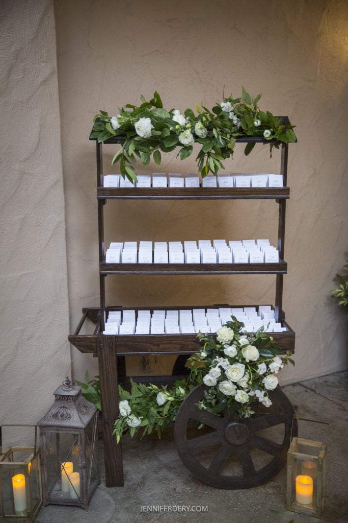 A rustic wooden cart with shelves stands against a beige wall, adorned with green leaves and white flowers, reminiscent of the serene ambiance captured at Rancho Bernardo Inn as wedding photos. The shelves are lined with small white cards, and two lanterns with candles are placed near the cart.