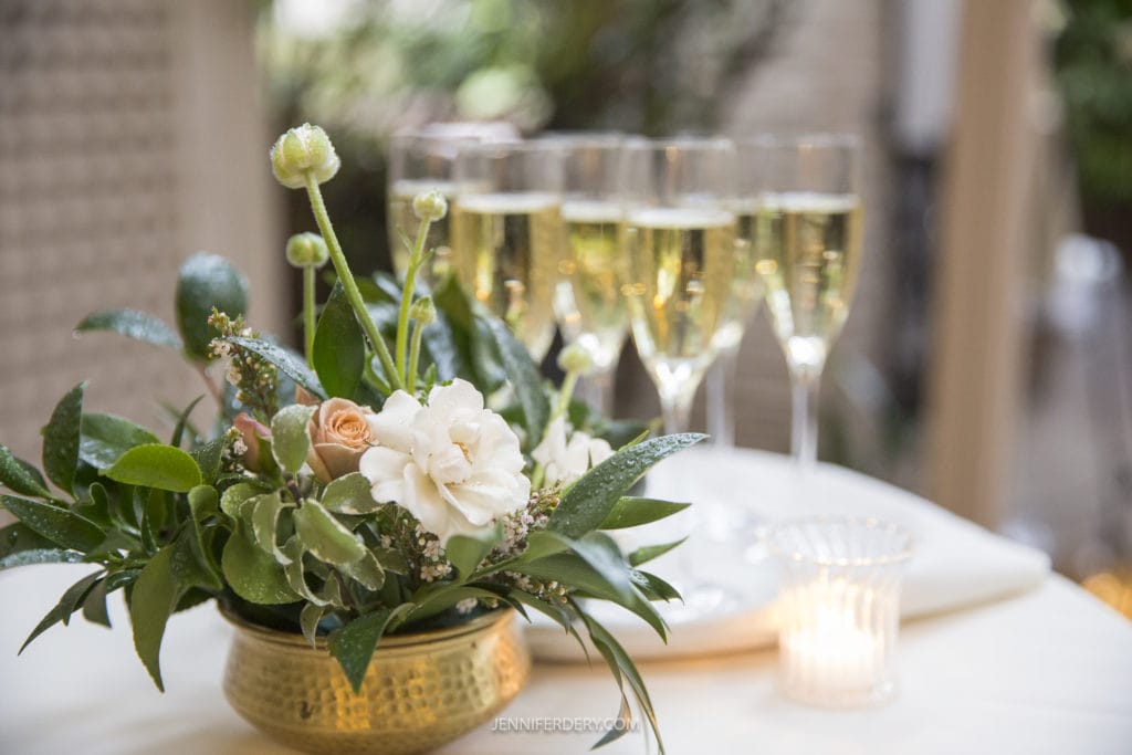 A close-up of a floral arrangement in a golden vase, featuring white and light peach roses, greenery, and white ranunculus flowers. In the background, five filled champagne glasses and a lit candle are visible on a blurred table, capturing the elegance typical of Rancho Bernardo Inn Wedding Photos.