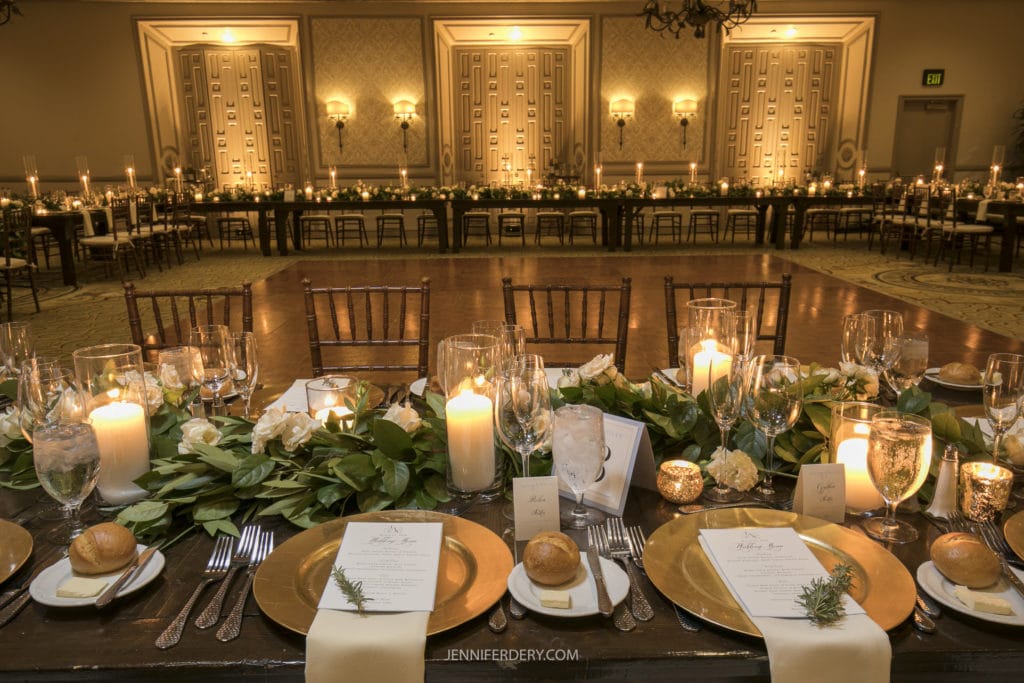 A beautifully set wedding reception table with elegant tableware, including gold chargers, white napkins, candles, and floral arrangements. Chairs are neatly lined up, and a larger, elaborately decorated table is visible in the background under soft lighting. Perfect for Rancho Bernardo Inn Wedding Photos.
