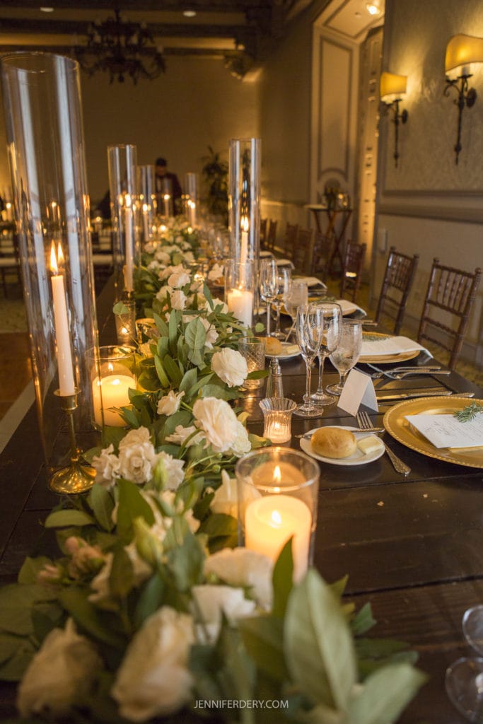 A long banquet table set for an elegant event, adorned with white roses, lush green garlands, and glowing candles in tall glass holders. The table features neatly arranged plates, glassware, and bread rolls on gold chargers, with warm ambient lighting enhancing the atmosphere—perfect for memorable Rancho Bernardo Inn wedding photos.