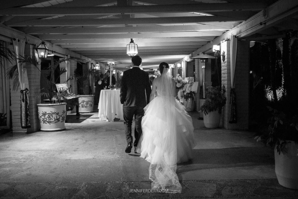 A newlywed couple walks hand-in-hand down a covered, well-decorated corridor at night. The bride wears an elegant, flowing gown, and the groom is in a suit. Captured from behind in their Founders Chapel wedding photos, they are illuminated by soft lights, adding a romantic atmosphere to the scene.