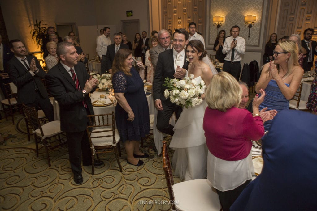 A bride dressed in white, holding a bouquet of flowers, walks down an aisle with a man in a suit, likely her father, while guests stand, applaud, and smile. The elegantly decorated reception hall captures the joy and celebration of this Founders Chapel wedding.