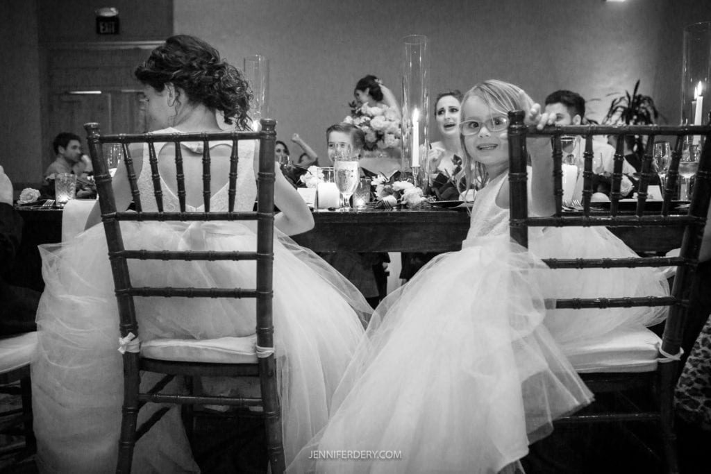 A black-and-white photo of two children in formal attire sitting on wooden chairs at a decorated table, facing away from the camera. One child turns around, smiling broadly and wearing glasses. Other wedding guests can be seen in the background of these charming Founders Chapel Wedding Photos.