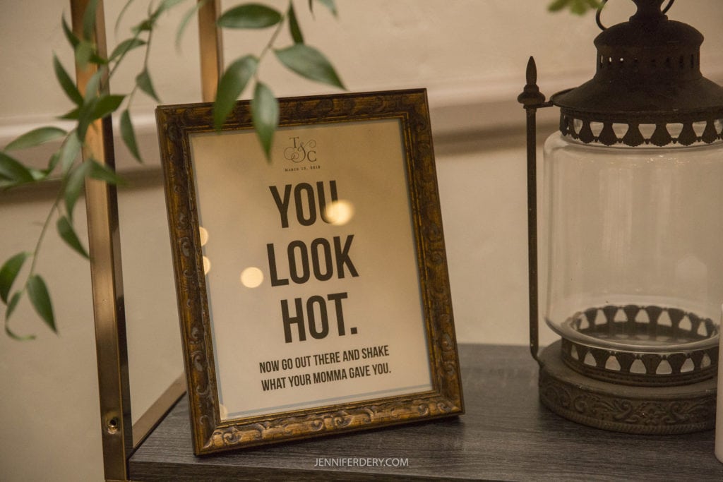 A framed sign on a tabletop reads, "YOU LOOK HOT. NOW GO OUT THERE AND SHAKE WHAT YOUR MOMMA GAVE YOU." The ornate frame sits beside a decorative lantern, and some green leaves from a plant are visible in the foreground. This charming scene would be perfect for Founders Chapel wedding photos.