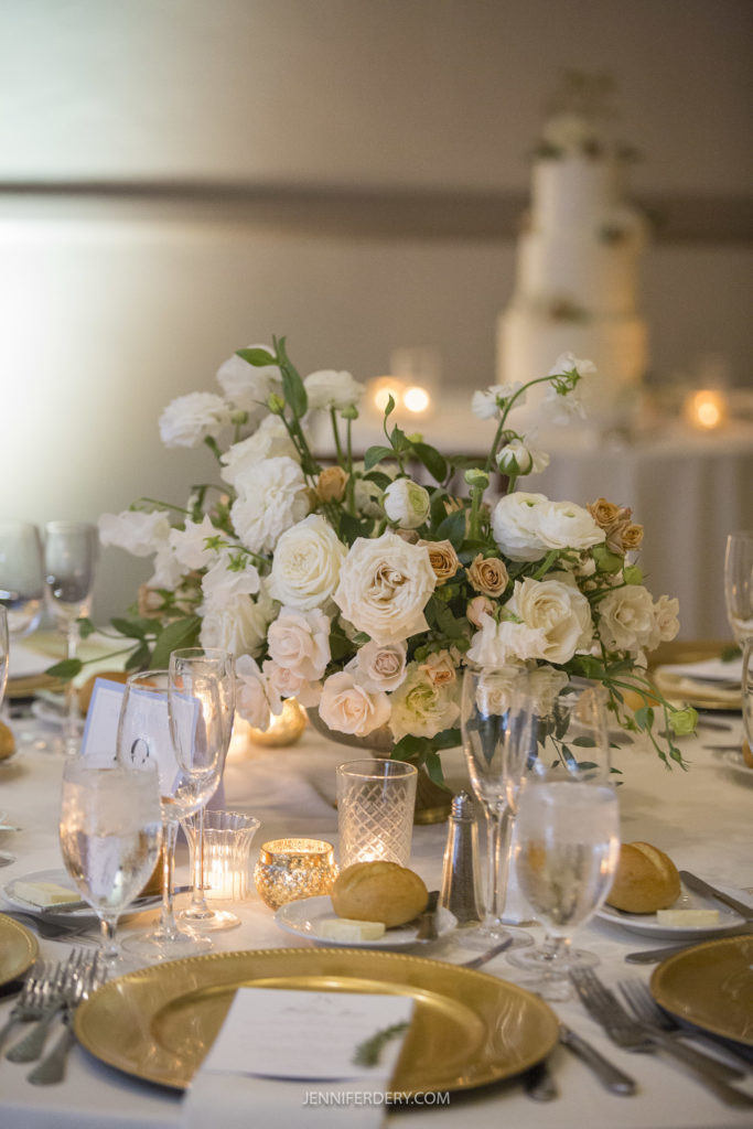 An elegant wedding table setting at Founders Chapel, featuring a centerpiece with white roses and greenery, surrounded by glasses, silverware, and plates with bread rolls on gold chargers. In the background, a multi-tiered white wedding cake is visible. Candles add a warm glow.