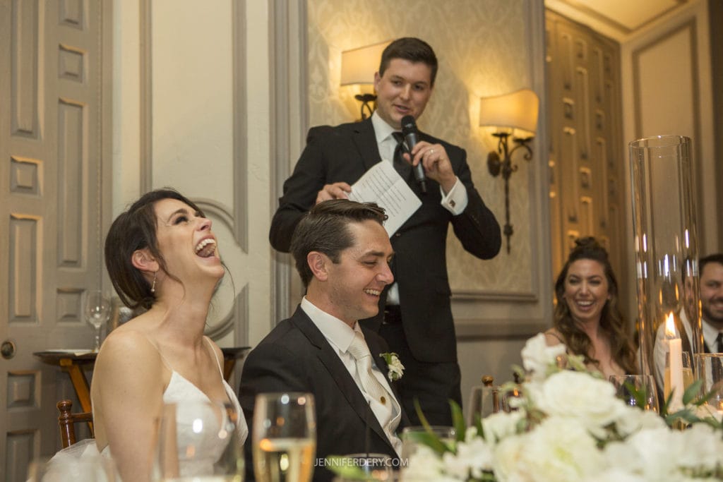 A bride and groom sit at a dinner table, laughing as a man in a suit gives a speech with a microphone. Two bridesmaids can be seen smiling in the background. The room, part of the Founders Chapel Wedding Photos collection, is elegantly decorated with flowers and candles on the table.