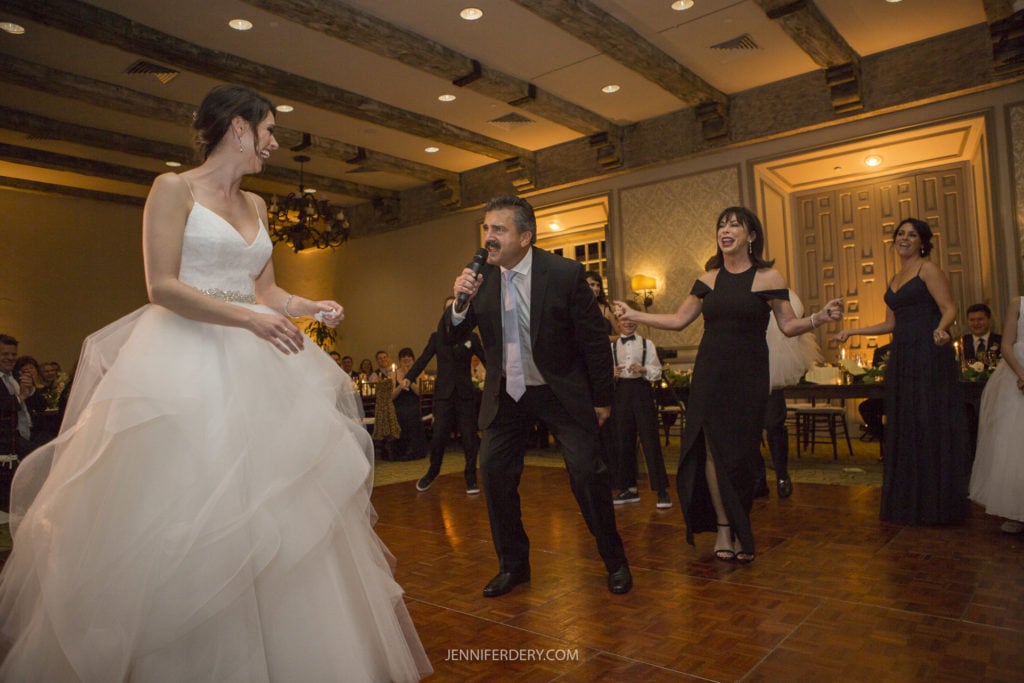 Bride in a white gown and her father in a suit with a microphone dance enthusiastically at their Founders Chapel Wedding Reception, joined by three women in black dresses. Guests are seated at tables in the background, enjoying the lively atmosphere and capturing priceless wedding photos.