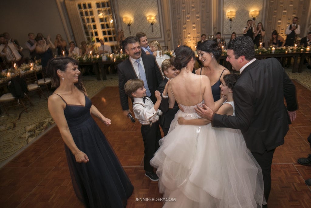 A bride in a white dress shares a joyful moment on the dance floor with her groom, three children in formal attire, and several well-dressed guests. Surrounded by onlookers and a beautifully set dining area with candlelit tables, this Founders Chapel Wedding Photos scene captures pure bliss.